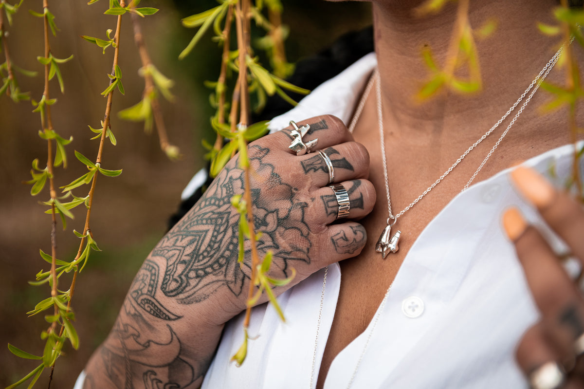 A photograph of a female models upper torso and one hand. She has tattoos on her arm, hands and fingers, wears a white shirt and alternative silver jewellery. She wears a sterling silver fox tooth ring, some simple silver stacking rings, a wider birch-bark style ring and a solid silver fox tooth on a silver chain.