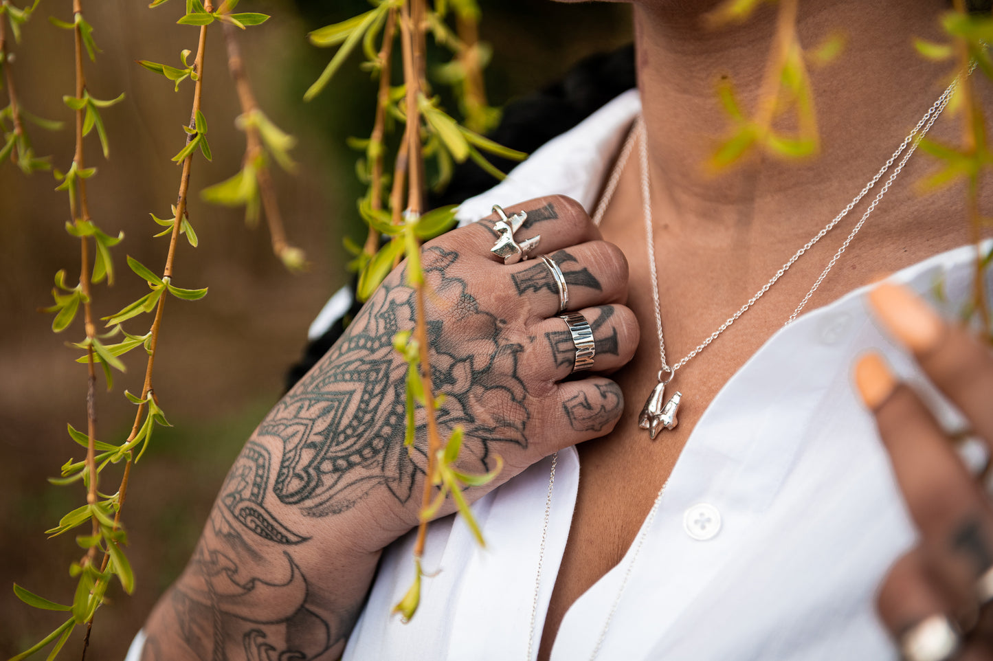 A close up photograph of a womans hand and upper torso amongst hanging vines. She wears a fox tooth necklace in sterling silver and an assortment of silver rings on her tattooed hand. On her forefinger she wears a sterling silver fox tooth ring.