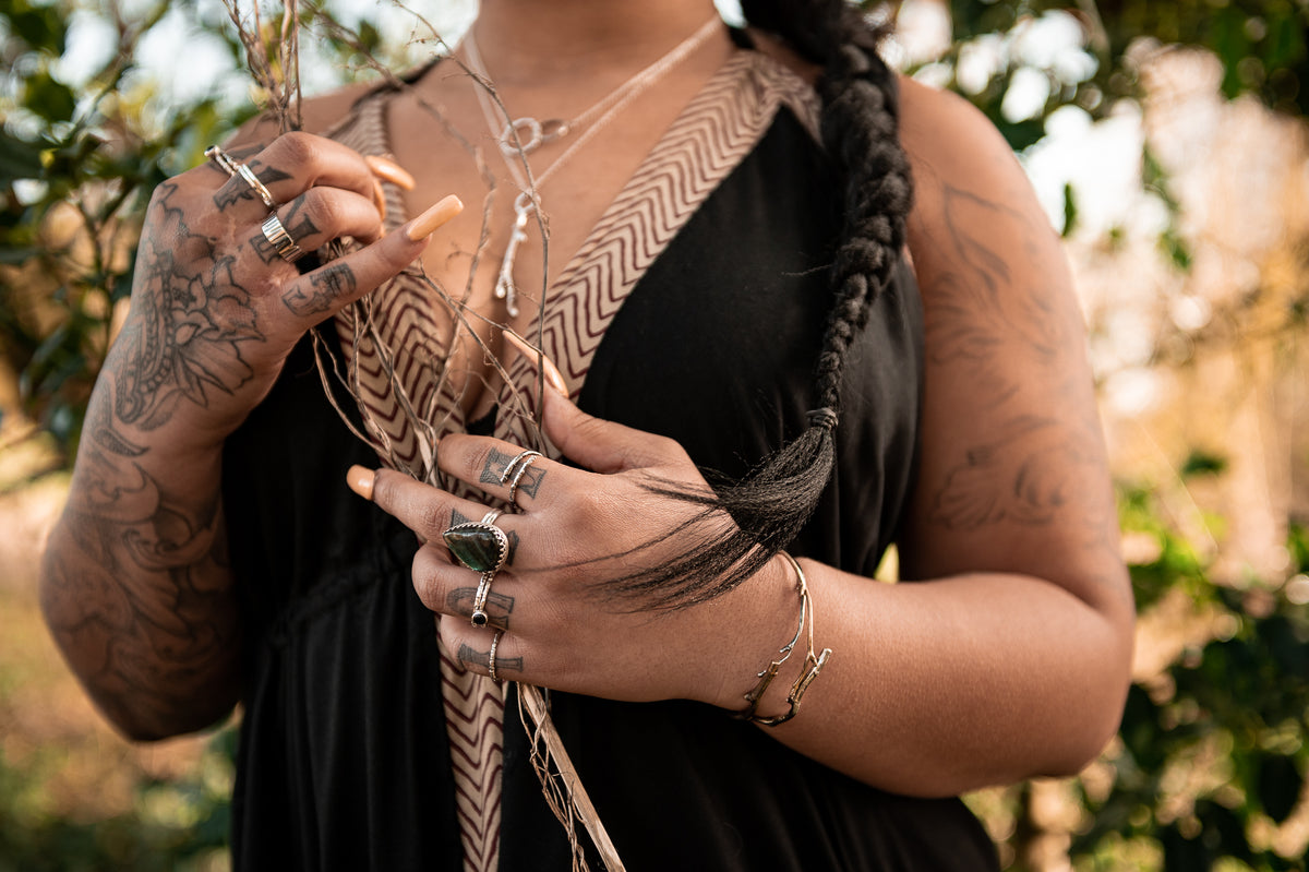 A black woman in witchy bohemian styled clothing poses in the woods. The shot is of her hands, arms and upper torso. She wears organically textured sterling silver and magical gemstone rings, bracelets, necklaces and pendants. The jewellery holds lots of luscious texture from twigs, bark and branches. The model has floral tattoos on her arms and hands.