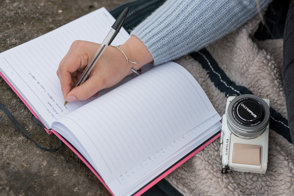 A girl sits outdoors on a rock, writing in a journal. She wears a cosy, blue sweater and a camera is sitting beside her notebook. Around her wrist, she wears a solid silver woodland bangle, that is made from a solid silver casting of a twig, gently wrapped around.