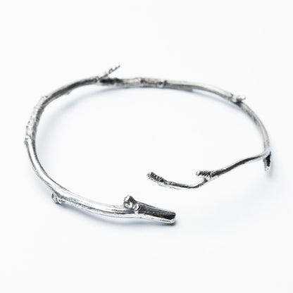 A silver bangle bracelet with a rough, natural texture, displayed on a white background. The bracelet is made from a piece of twig that has been cast in solid sterling silver.